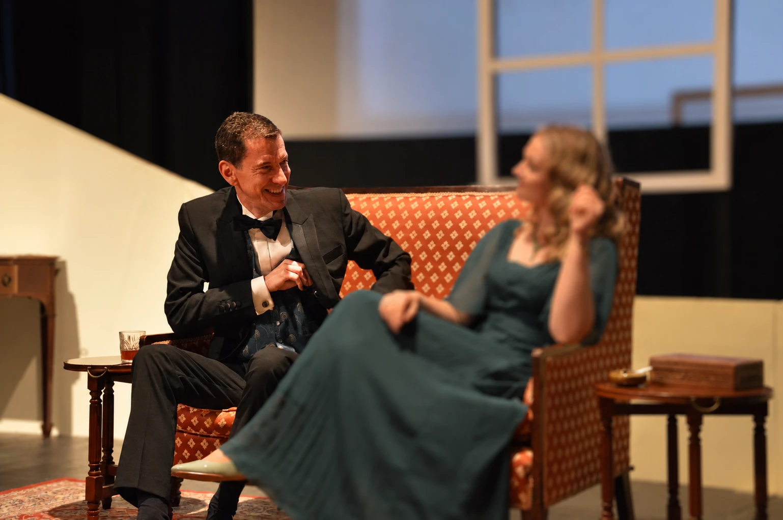 Paris seated on a sofa in a Noël Coward comedy, alongside another performer whose face is obscured